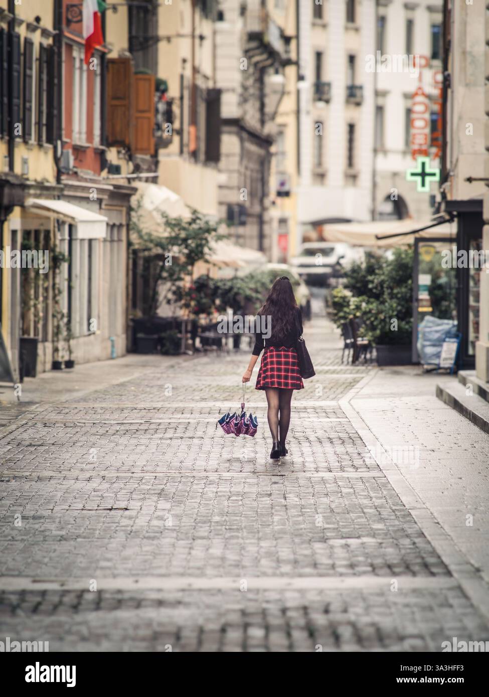 Une jeune femme élégante dans une jupe à carreaux rouge descend une rue pavée italienne tenant un parapluie. L'architecture historique et l'éclairage doux c Banque D'Images