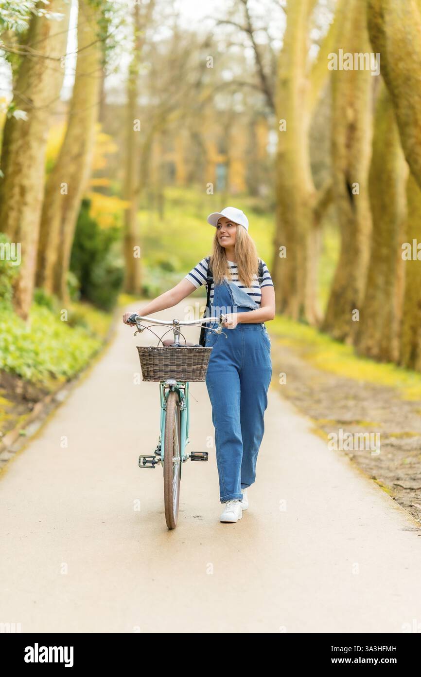 Heureuse jeune femme marchant avec son vélo vintage dans un parc, profitant d'une journée ensoleillée de loisirs Banque D'Images