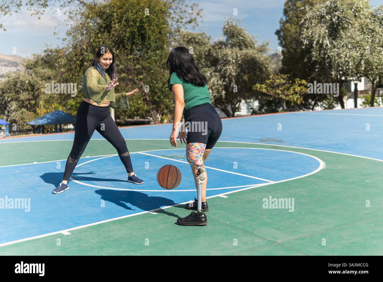 Deux jeunes femmes jouant au basket-ball par une journée ensoleillée, dont une avec une prothèse de jambe montrant détermination et inclusivité dans le sport Banque D'Images