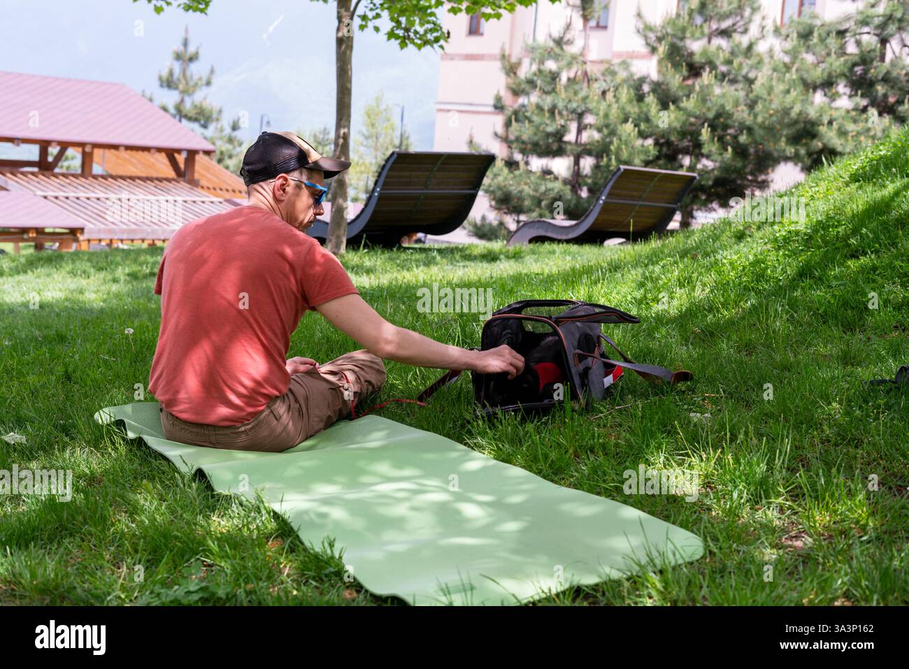 L'homme en T-shirt rouge est assis sur un tapis de yoga vert dans un parc verdoyant herbeux, caressant doucement le chat noir à l'intérieur d'un transporteur portable pour animaux de compagnie. Le chat porte un harnais rouge, peekin Banque D'Images