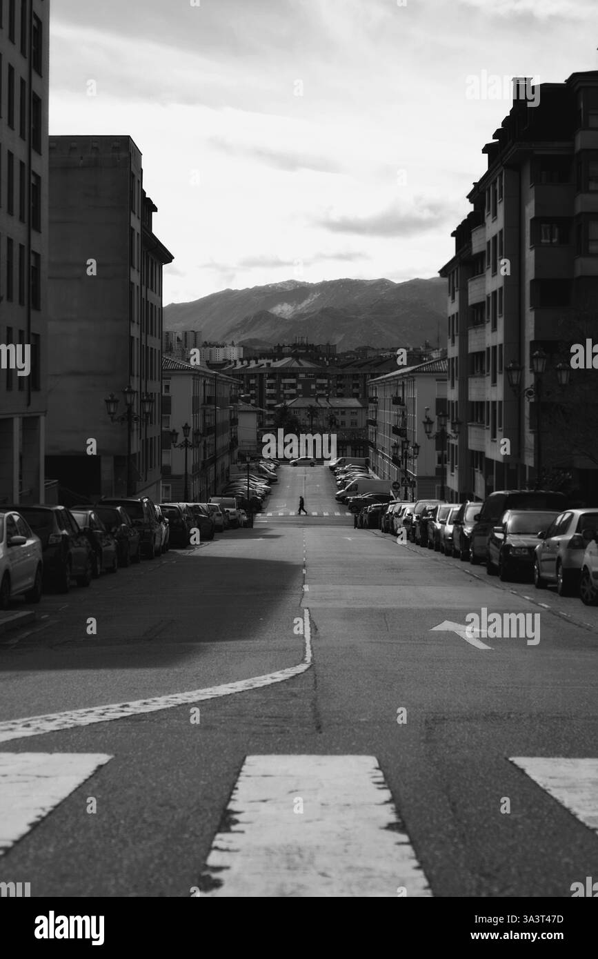 Photo en noir et blanc d'une rue urbaine avec des voitures garées, des montagnes lointaines et un piéton traversant la route sous un ciel nuageux Banque D'Images