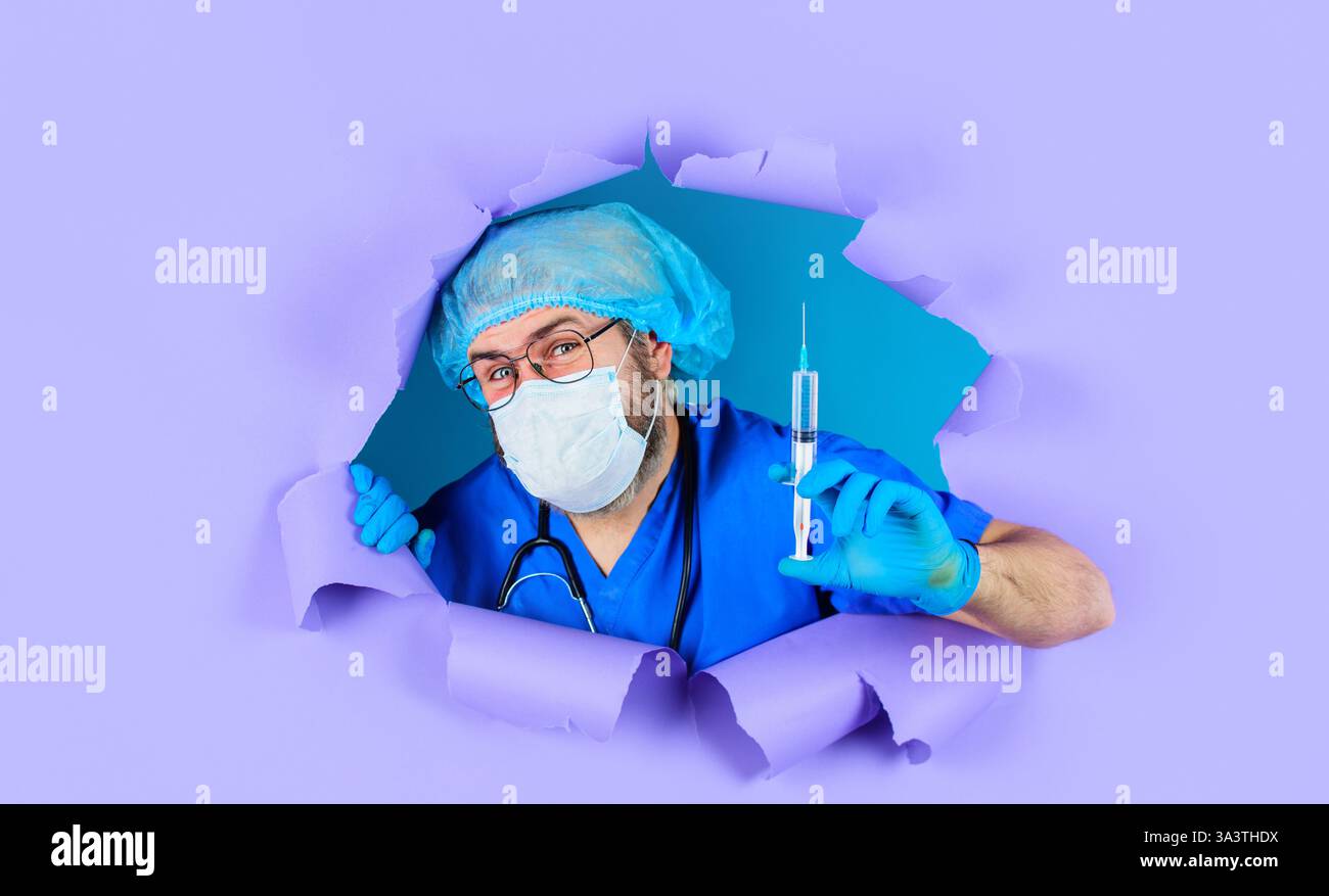 Docteur en masque médical et capuchon chirurgical avec seringue regardant à travers le trou dans le papier. Infirmier ou médecin en uniforme et gants en caoutchouc tenant la syrine Banque D'Images
