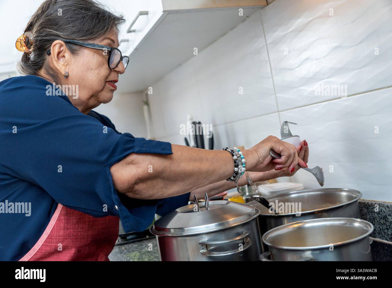 Femme senior latine portant un tablier ajoutant des ingrédients à une soupe dans sa cuisine à la maison Banque D'Images
