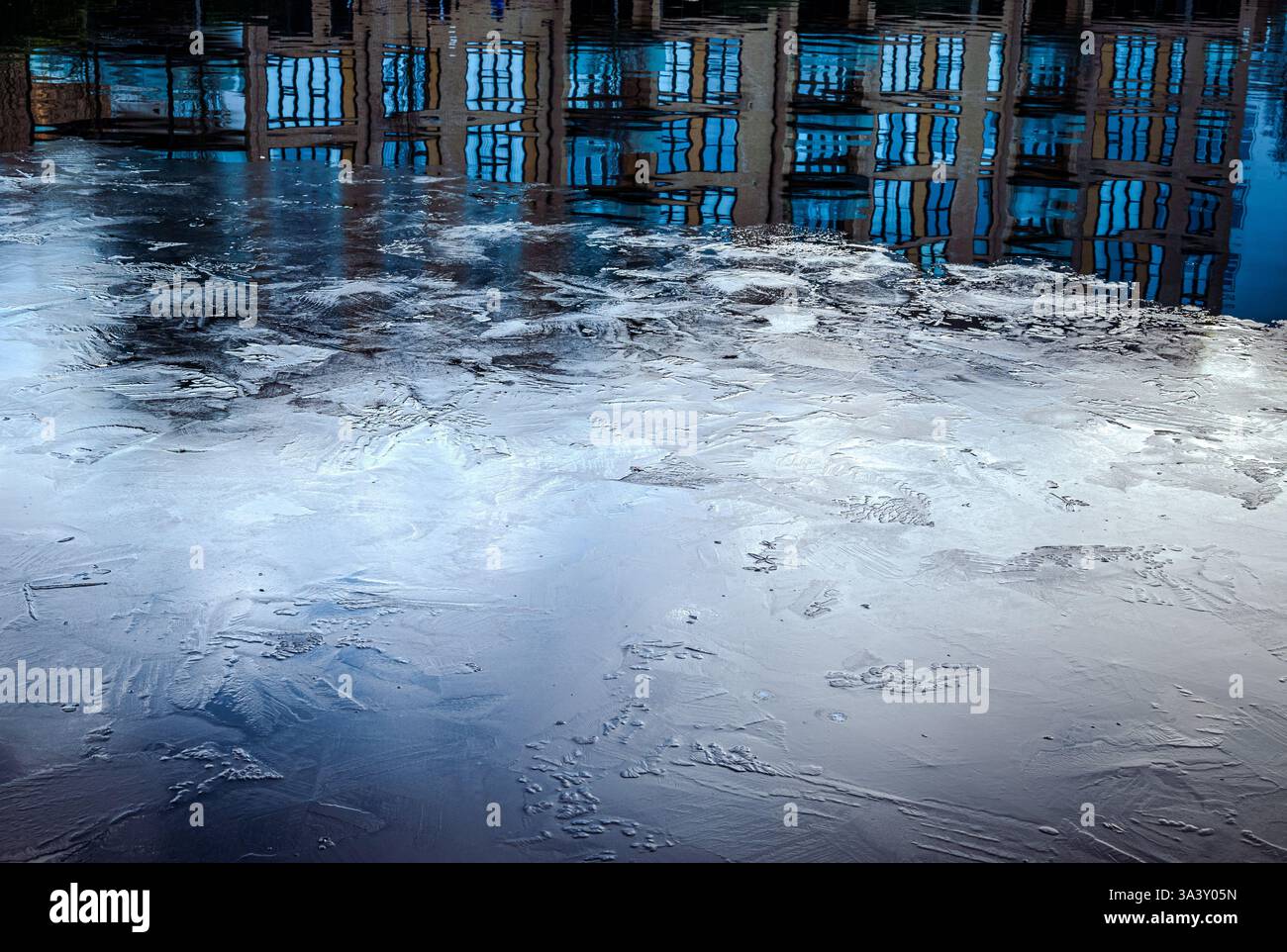 La surface de l’eau gelée est gravée de rayures abstraites et de textures, tandis qu’une section partiellement non gelée reflète les immeubles d’habitation Banque D'Images
