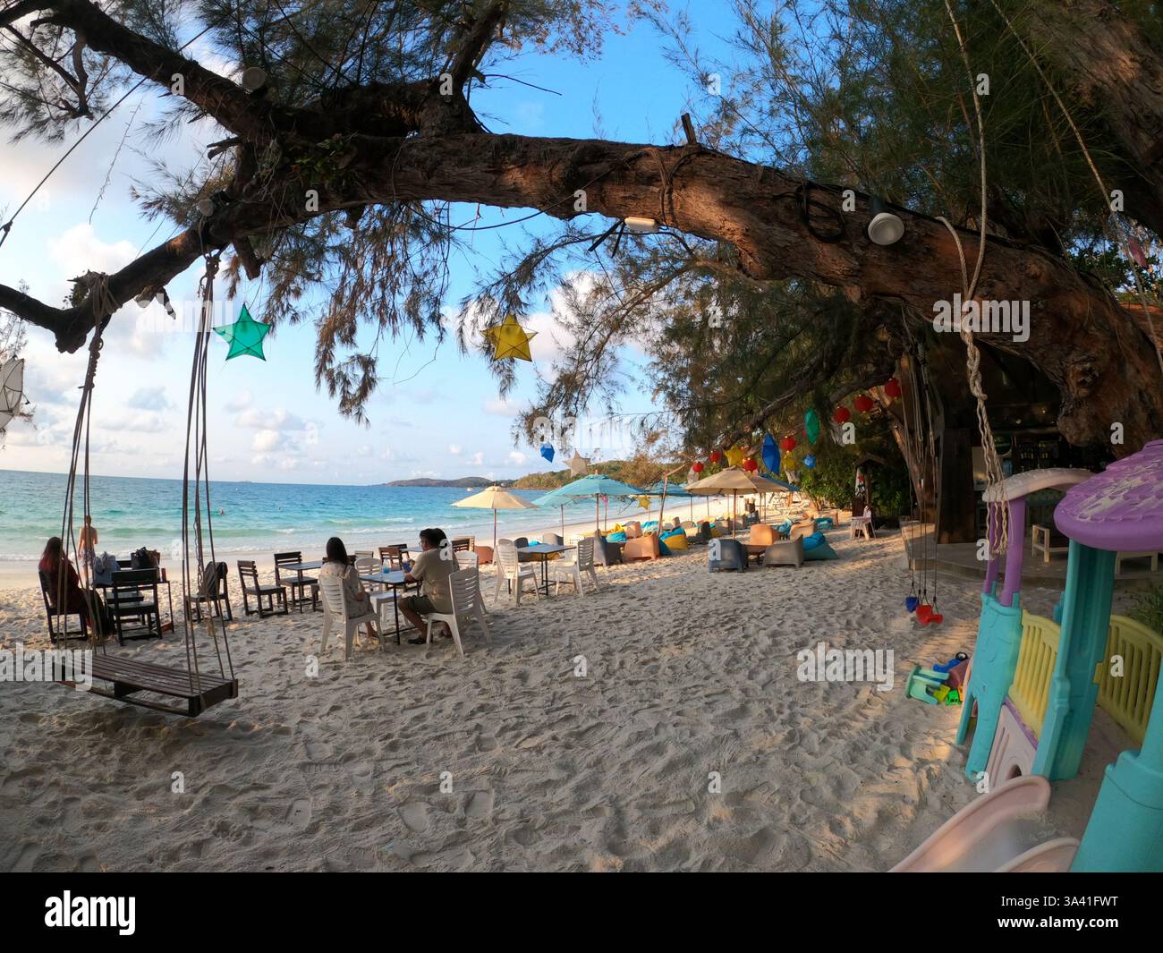Un café paisible en bord de mer à l'ombre des arbres, où les gens se détendent au bord de l'océan. Balançoires, lumières et sable doux créent une ambiance tropicale parfaite. Banque D'Images