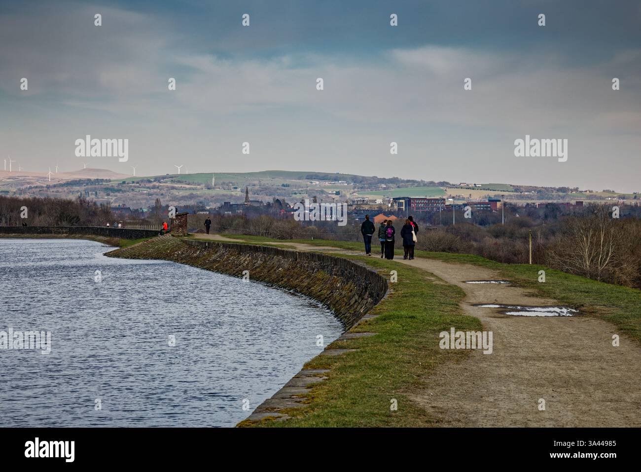 Un groupe familial se promène le long d'un chemin sur le mur du barrage d'Elton Reservoir avec la ville de Bury au loin, Greater Manchester. Banque D'Images