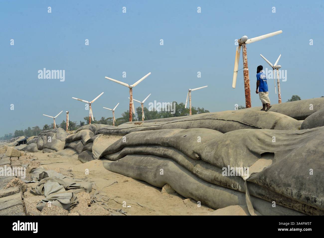 Un homme du coin marche près des éoliennes sur les rives de l’île de Kutubdia. Autrefois symbole d’espoir pour l’énergie verte, ces structures rouillées sont aujourd’hui abandonnées. Une fois opérationnels et produisant de l’électricité, ils sont tombés en délabrement, suscitant des inquiétudes quant à la mauvaise gestion et à l’avenir des initiatives du Bangladesh en matière d’énergies renouvelables. Île de Kutubdia, Bangladesh. Banque D'Images