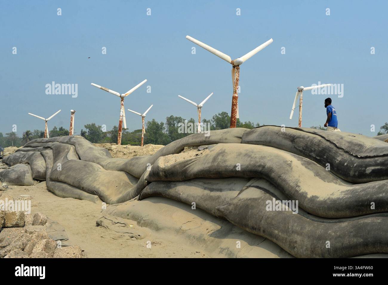 Un homme du coin marche près des éoliennes sur les rives de l’île de Kutubdia. Autrefois symbole d’espoir pour l’énergie verte, ces structures rouillées sont aujourd’hui abandonnées. Une fois opérationnels et produisant de l’électricité, ils sont tombés en délabrement, suscitant des inquiétudes quant à la mauvaise gestion et à l’avenir des initiatives du Bangladesh en matière d’énergies renouvelables. Île de Kutubdia, Bangladesh. Banque D'Images