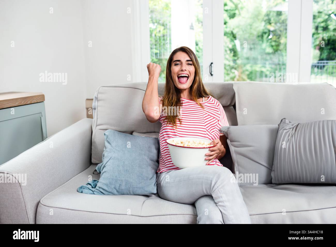 Femme excitée sur le canapé mangeant du pop-corn et acclamant tout en regardant la télévision Banque D'Images