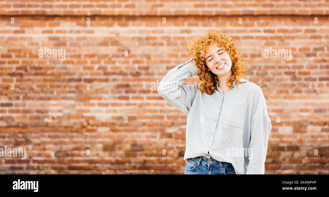 Portrait d'une jeune femme joyeuse aux cheveux roux bouclés, portant une chemise gris clair et un Jean, touchant ses cheveux et souriant les yeux fermés, debout Banque D'Images Portrait d'une jeune femme joyeuse aux cheveux roux bouclés, portant une chemise gris clair et un Jean, touchant ses cheveux et souriant les yeux fermés, debout Banque D'Images