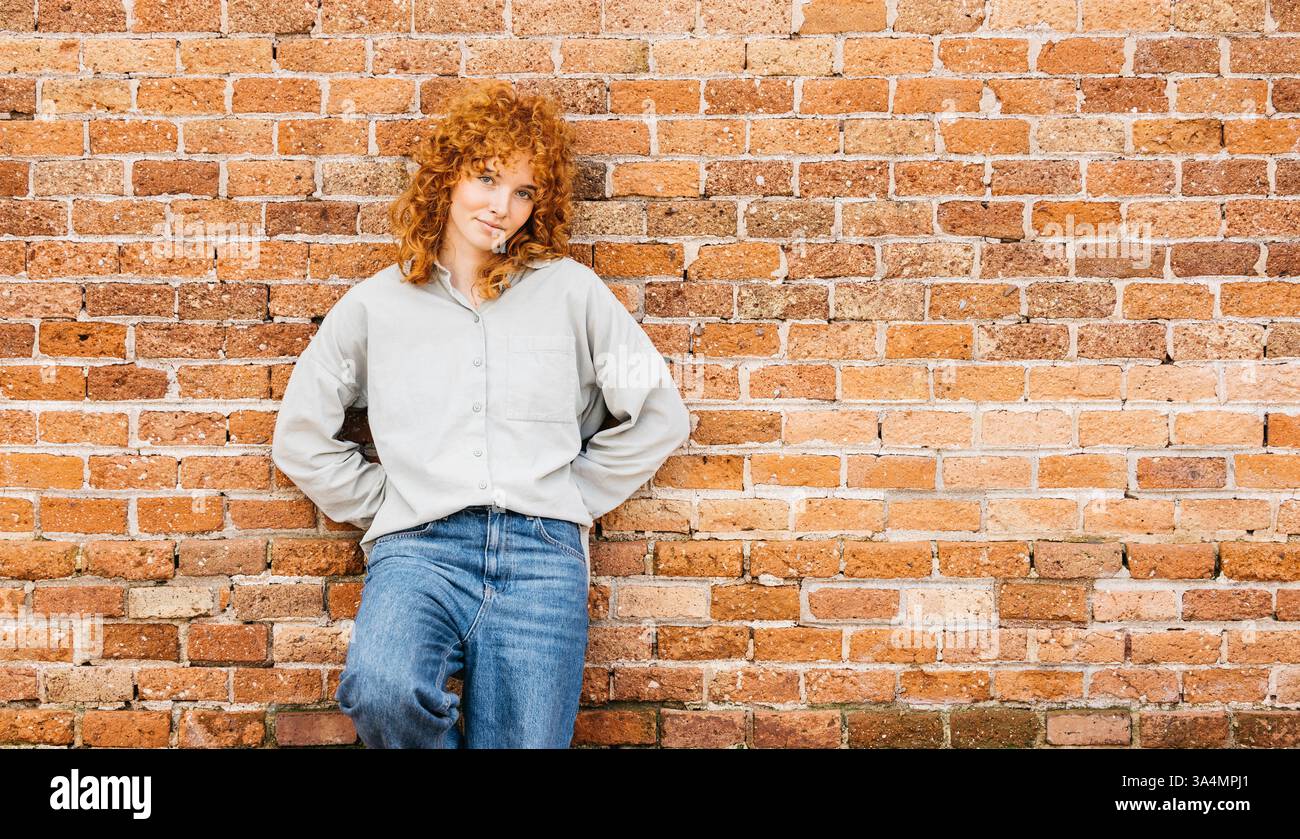 Jeune femme décontractée avec les cheveux roux bouclés appuyée contre un mur de briques Banque D'Images Jeune femme décontractée avec les cheveux roux bouclés appuyée contre un mur de briques Banque D'Images