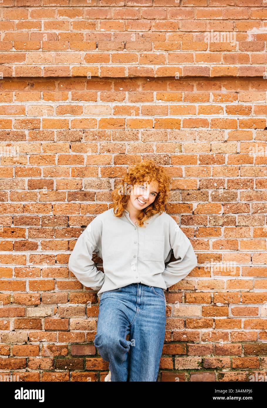 Portrait d'une jeune femme souriante avec des cheveux roux bouclés appuyés contre un mur de briques, regardant la caméra Banque D'Images Portrait d'une jeune femme souriante avec des cheveux roux bouclés appuyés contre un mur de briques, regardant la caméra Banque D'Images