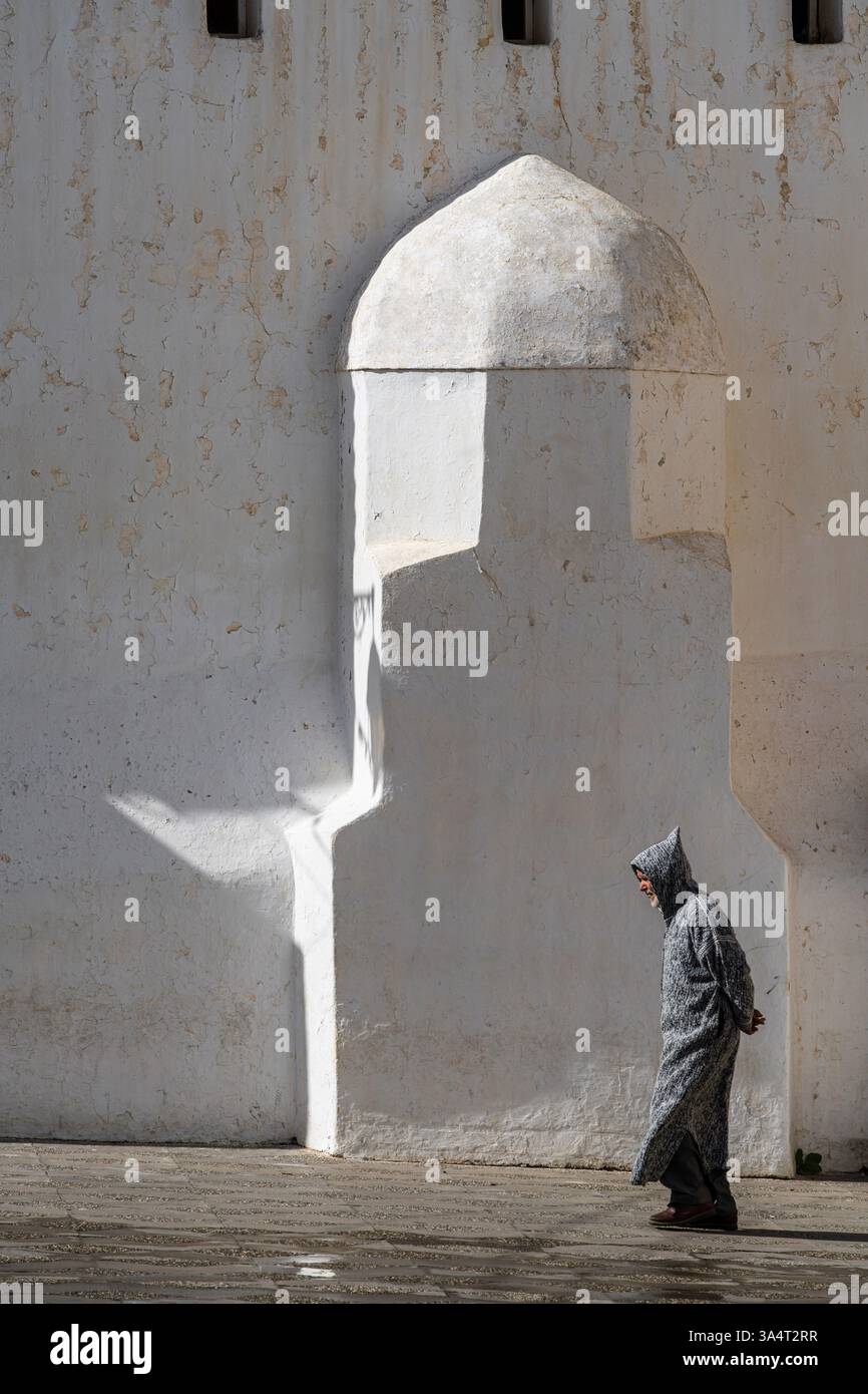 Hommes solitaires dans les djellabas d'hiver passant par la mosquée Saida, place Abdallah Guennoun, Asilah, Maroc, Afrique du Nord Banque D'Images