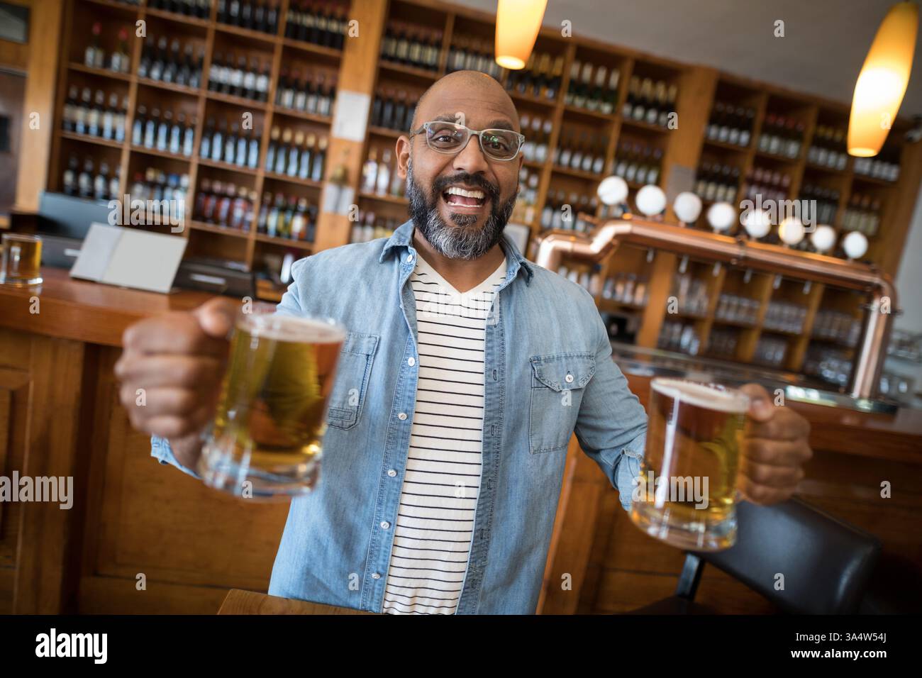 Homme souriant tenant des tasses à bière dans un bar animé avec des étagères en bois Banque D'Images