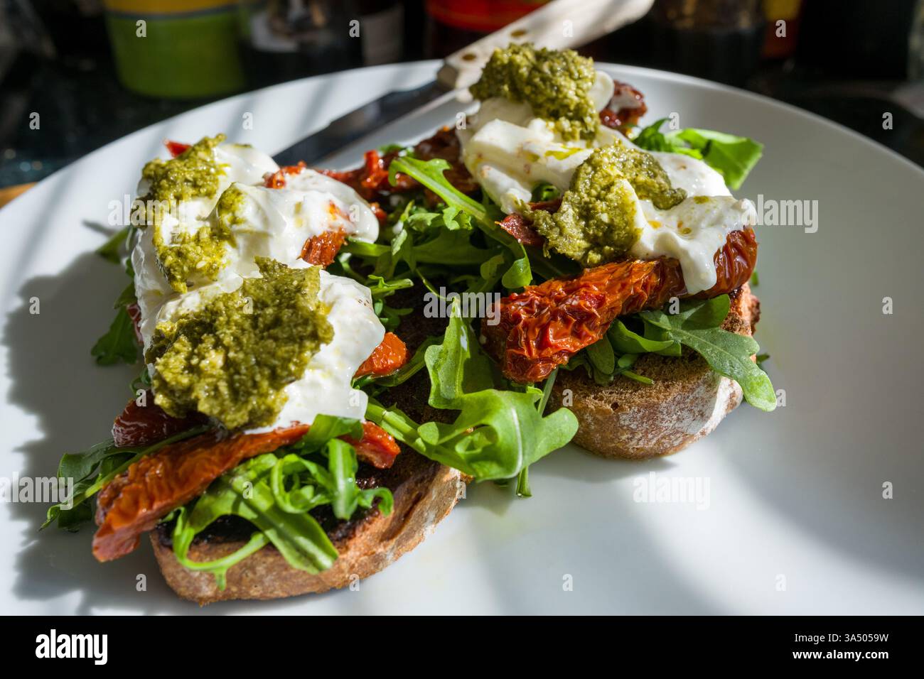 Seigle grillé et levain à l'épeautre arrosé d'huile d'olive et recouvert de roquette, de tomates séchées, de burrata crémeuse et de pesto au basilic. Un t méditerranéen Banque D'Images
