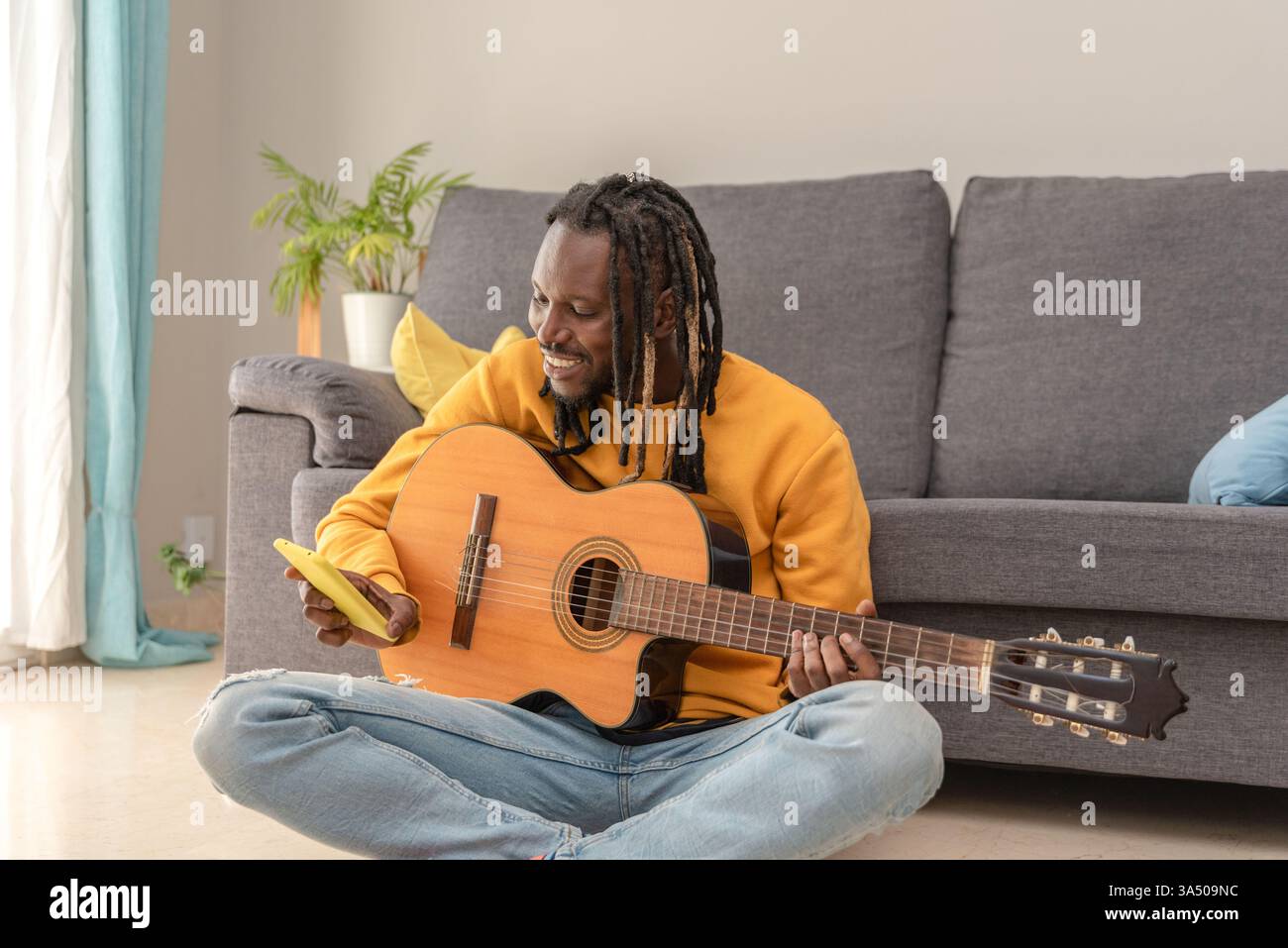 Musicien masculin noir avec dreadlocks à l'aide d'un smartphone assis avec guitare sur le sol à la maison Banque D'Images