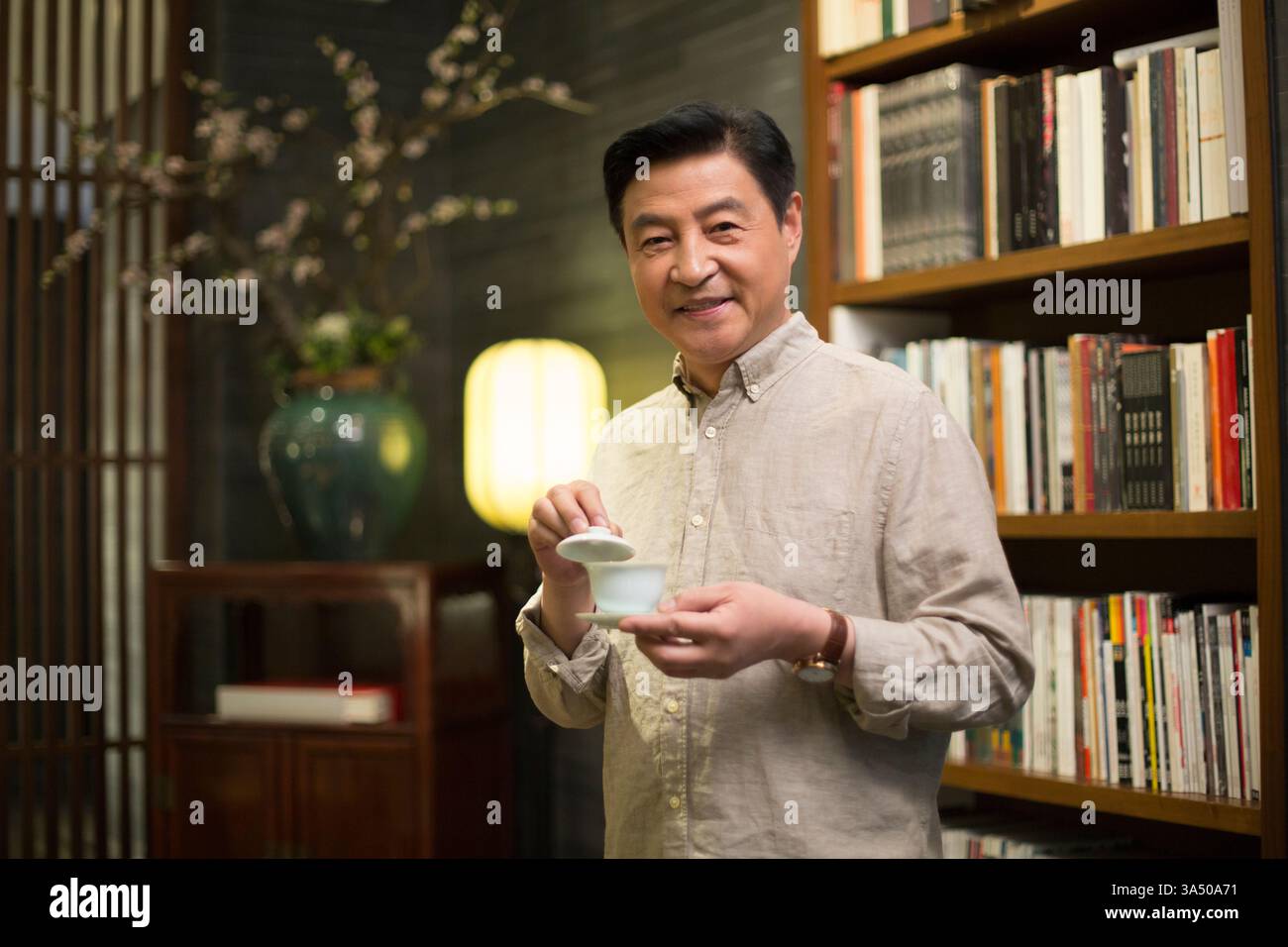 L'homme chinois souriant aime une tasse de thé à côté d'une étagère à la maison. Cette photo chaleureuse et de style de vie convient à la culture du thé, à la vie à la maison et aux thèmes de marque personnelle ou de petite entreprise. Banque D'Images