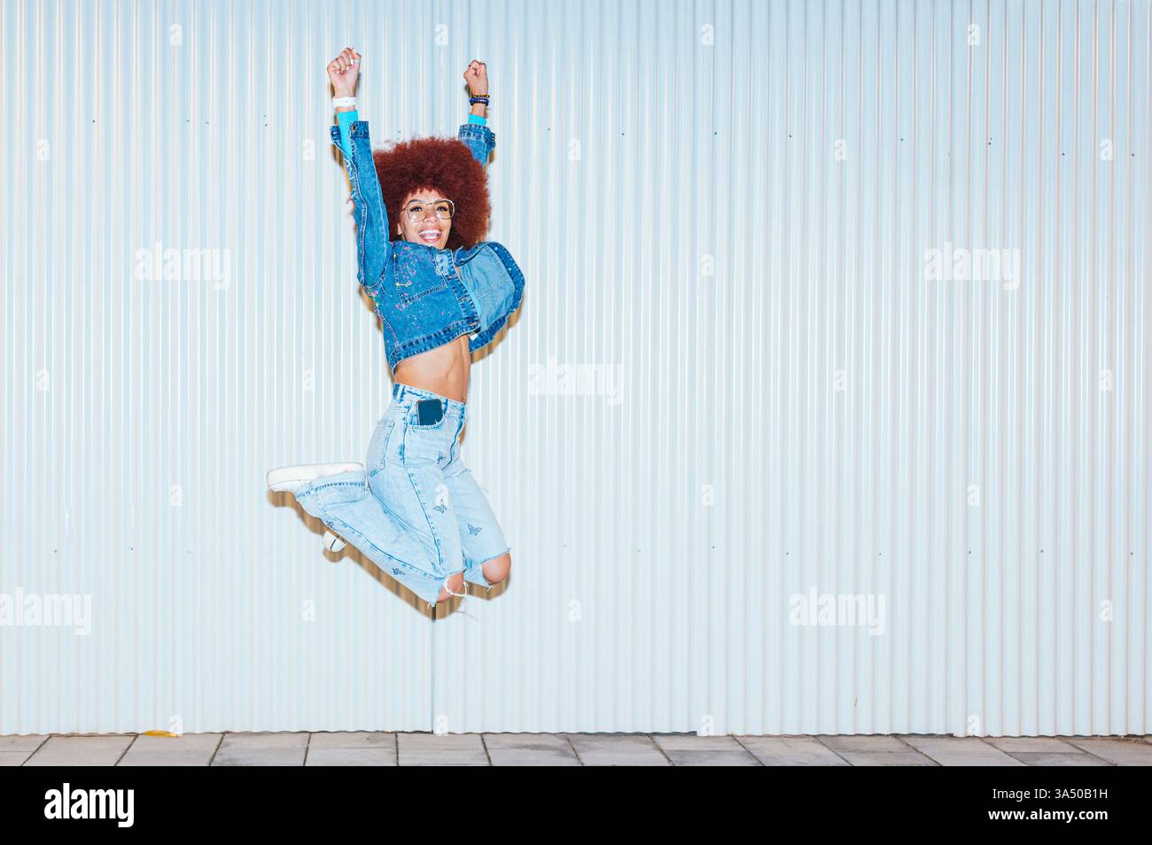 Femme noire avec une coiffure afro saute avec les bras levés contre un mur côtelé dans un plan de studio dynamique. Sa pose énergique et son expression lumineuse transmettent confiance en soi, positivité et style de vie actif. Parfait pour la mode, l'autonomisation ou les campagnes de bien-être. Banque D'Images
