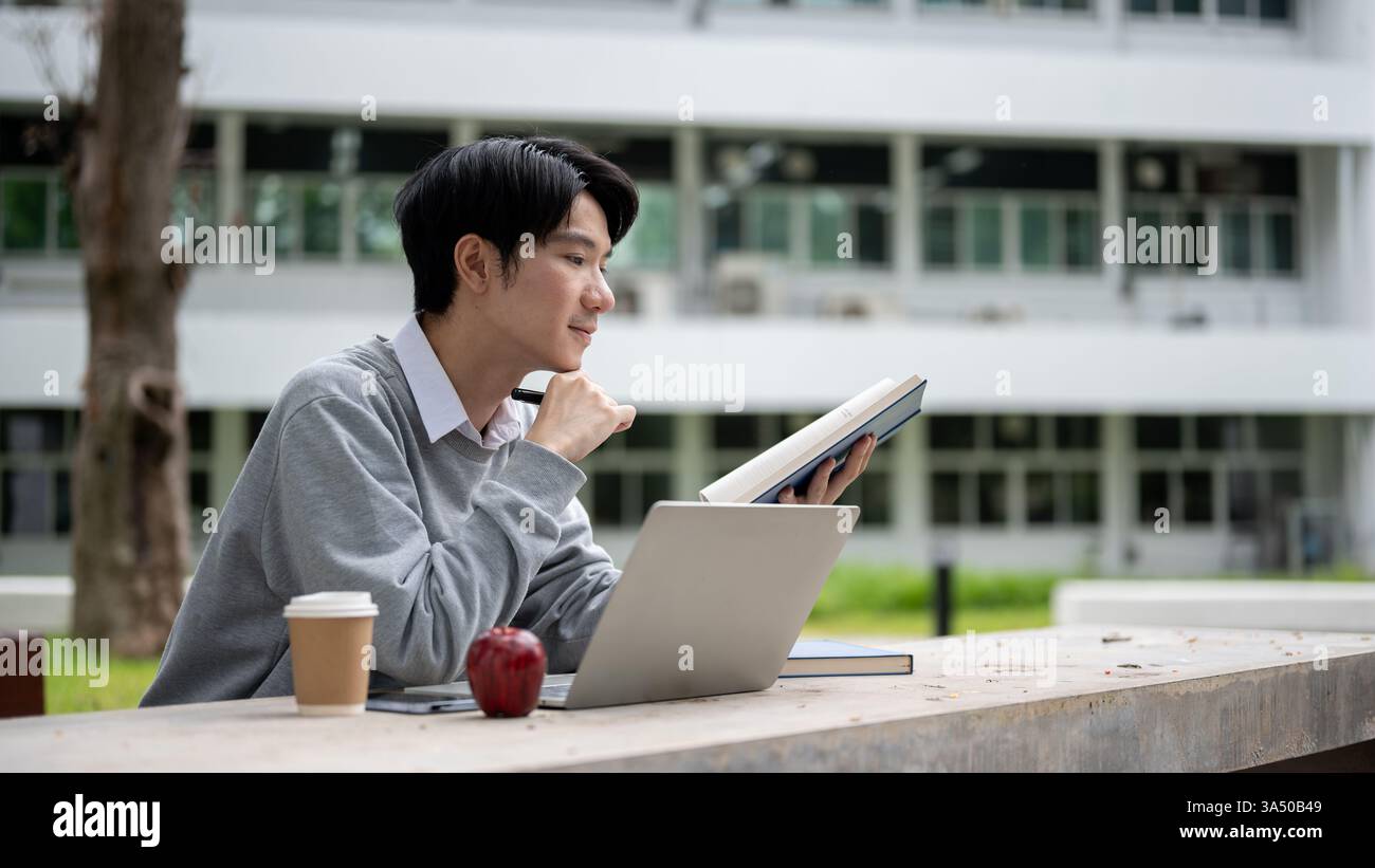 Étudiant masculin asiatique penchant le menton sur la main tout en tenant et lisant le livre assis avec des gadgets et des aliments à table sur le campus pendant la journée Banque D'Images