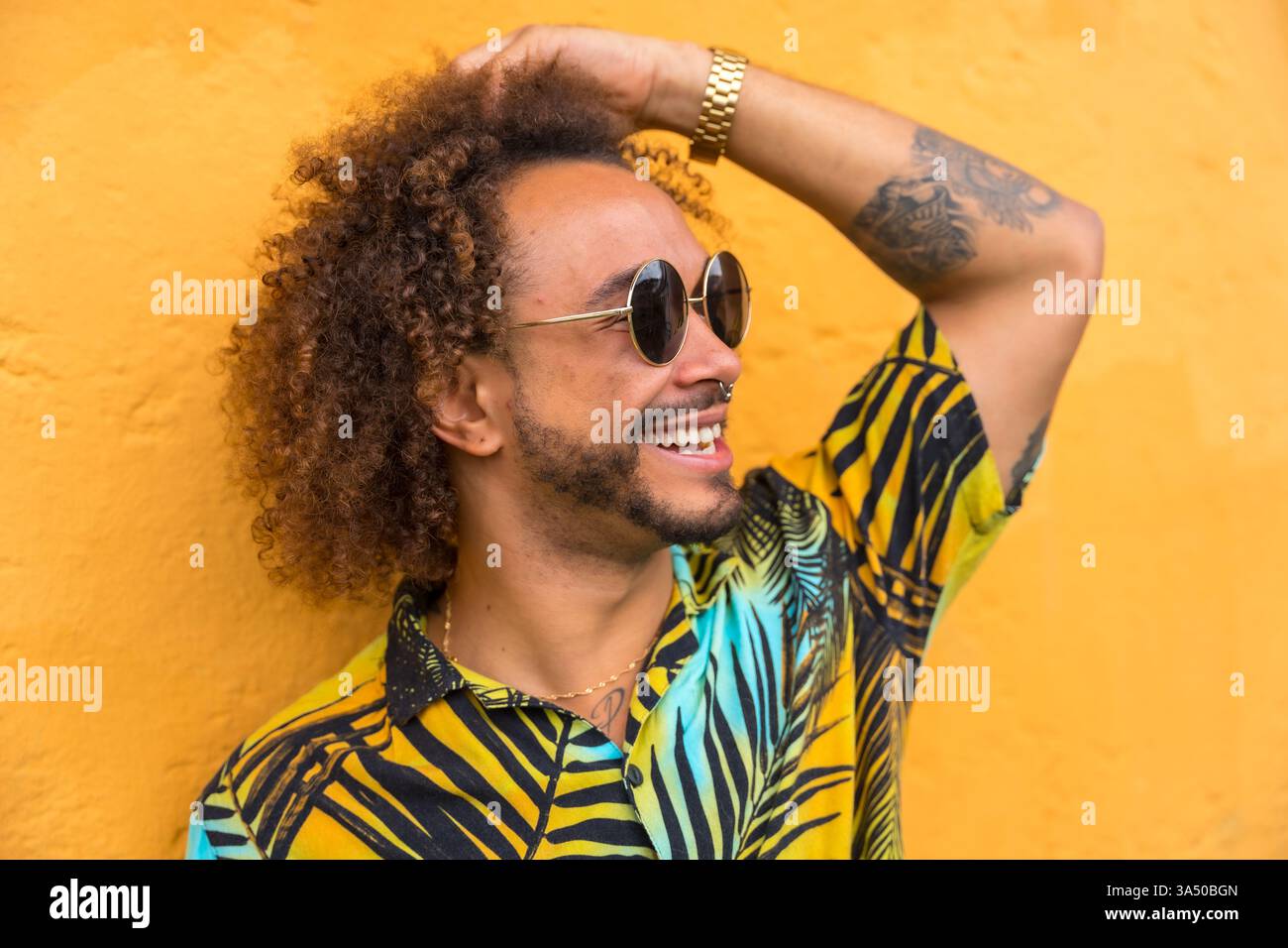 Un homme avec un Afro porte une chemise imprimée palmier sur un fond jaune vif. La couleur audacieuse et le style décontracté rendent ce portrait idéal pour les campagnes de mode et de style de vie. Utile pour la mode masculine, les portefeuilles de mannequins et le style de produit. Banque D'Images