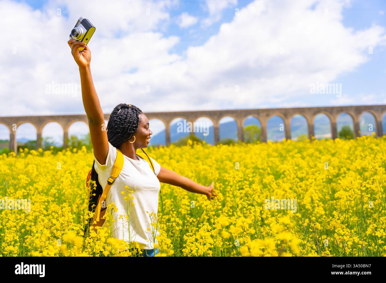Femme noire avec des tresses voyage à travers un champ de fleurs jaunes, capturant un moment de liberté avec un appareil photo. Scène printanière lumineuse mettant en valeur les voyages, la nature et l'aventure en plein air. Parfait pour le style de vie, la photographie et les thèmes d'exploration. Banque D'Images