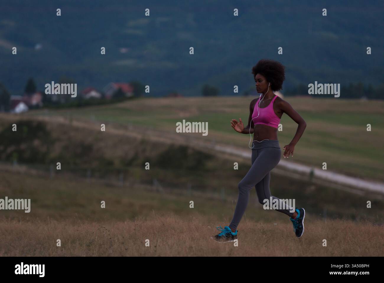 Image pleine longueur dynamique d'une jeune coureuse noire faisant du jogging à l'extérieur la nuit dans un cadre naturel. Elle porte des écouteurs, mettant en valeur la forme physique et un mode de vie sain. Parfait pour le sport, le bien-être, les activités de plein air et les campagnes de style de vie actif. Banque D'Images