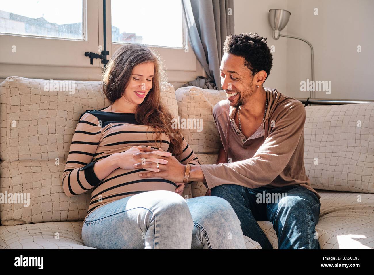 Portrait de style de vie chaleureux d'un couple multiethnique à la maison. Un homme noir touche doucement le ventre de sa femme enceinte alors qu'ils sont assis sur un canapé, souriant avec un bonheur attendu. Banque D'Images