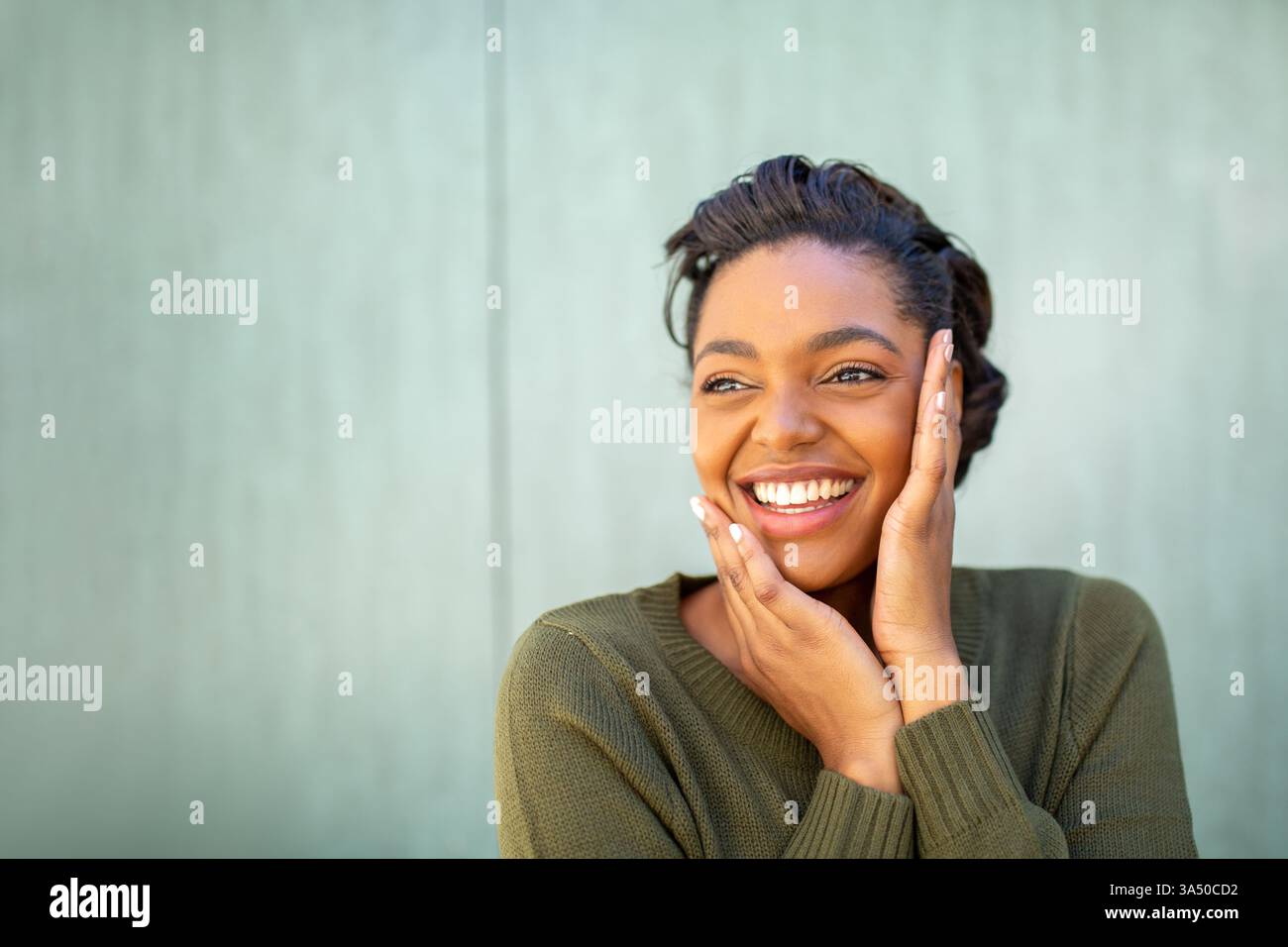 Portrait lumineux d'une femme noire souriante debout dehors contre un mur vert pendant la journée. Elle regarde loin avec les mains sur son visage, projetant une confiance détendue. Banque D'Images
