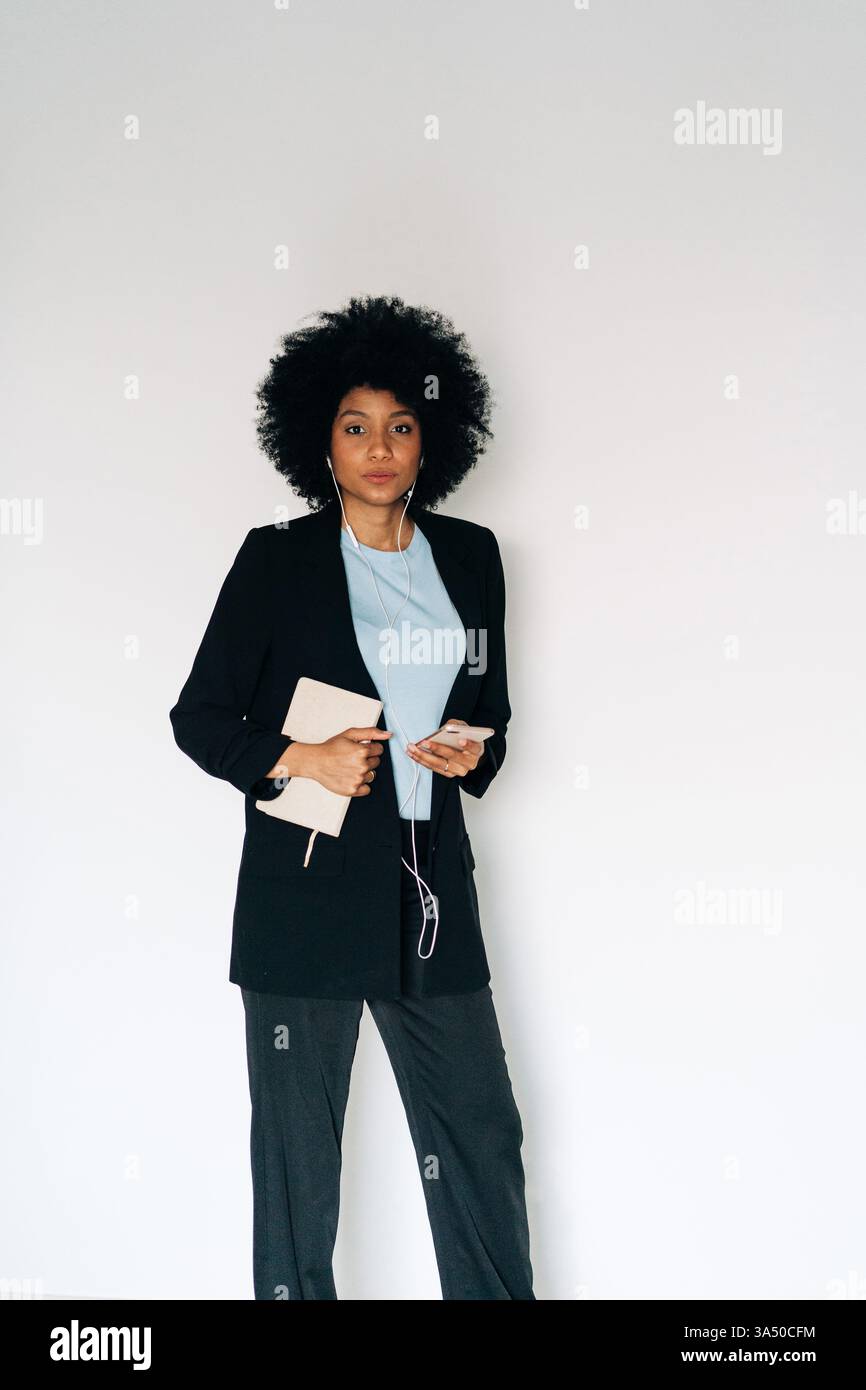 Femme hispanique avec un téléphone portable et des écouteurs debout sur un fond blanc. Le portrait fin et technophile met en valeur la connectivité contemporaine et le style personnel, idéal pour les applications, les appareils mobiles ou les campagnes technologiques. Banque D'Images