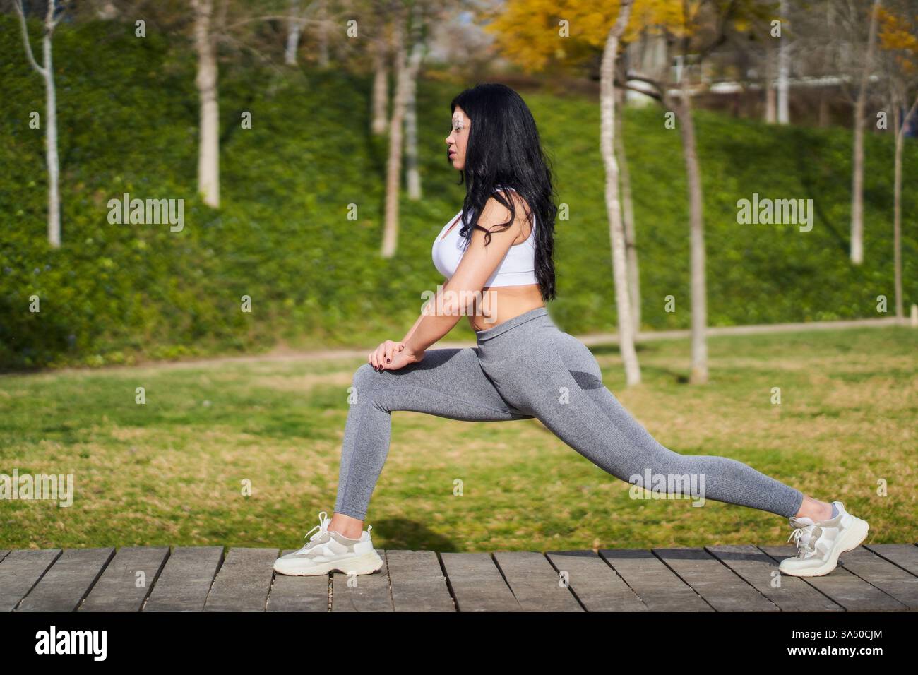 Vue de côté d'une jeune femme en tenue de sport exécutant des squats dans un parc ensoleillé. Idéal pour les campagnes de fitness et de bien-être, l'entraînement en plein air et les histoires de style de vie actif. Banque D'Images