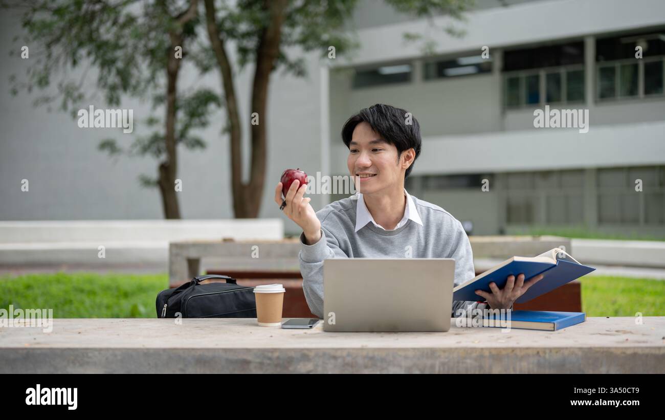 Étudiant masculin asiatique souriant assis avec un ordinateur portable à table tenant pomme et livre pendant l'heure de pause sur le campus pendant la journée Banque D'Images