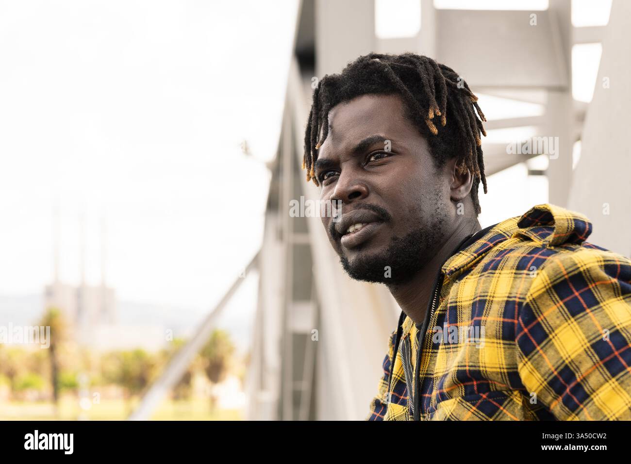 Homme africain assis dehors, appuyé contre une structure métallique et regardant loin. Le portrait décontracté et contemporain fonctionne pour le style de vie, la mode et les thèmes de marque personnelle. Convient aux campagnes axées sur la confiance et la vie urbaine. Banque D'Images