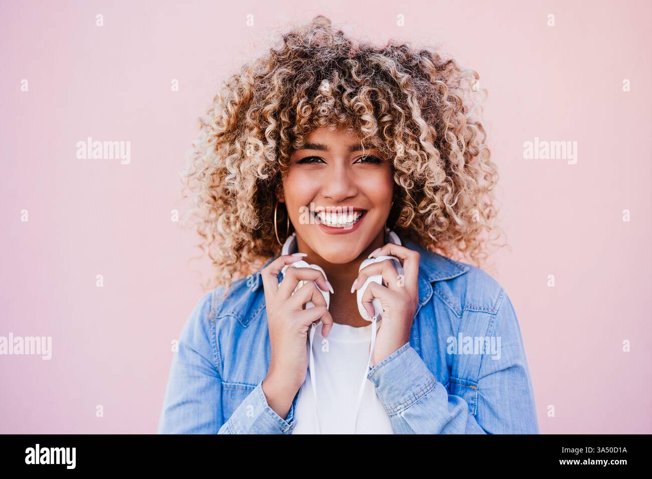 Femme hispanique souriante avec un afro bouclé porte des écouteurs autour du cou sur un fond rose. L'image transmet un moment de style de vie joyeux axé sur la musique et le style personnel. Idéal pour les thèmes de style de vie, de mode et de culture urbaine. Banque D'Images