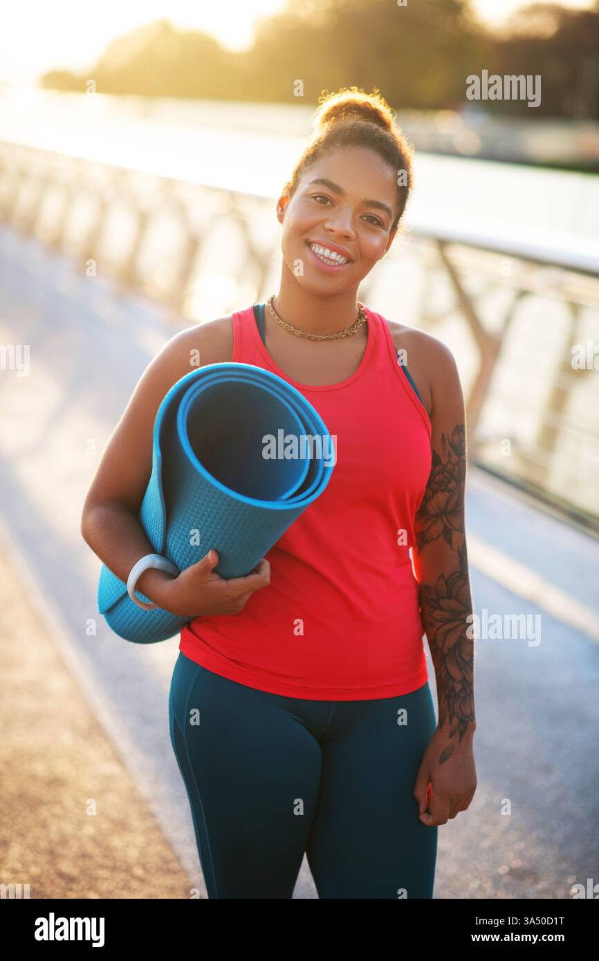 Portrait d'une femme confiante tenant un tapis de sport, prête pour l'entraînement. Elle rayonne de motivation et d'excitation pour une séance d'entraînement dans le cadre d'un mode de vie actif. Idéal pour les campagnes de fitness axées sur la préparation et l'énergie positive. Banque D'Images