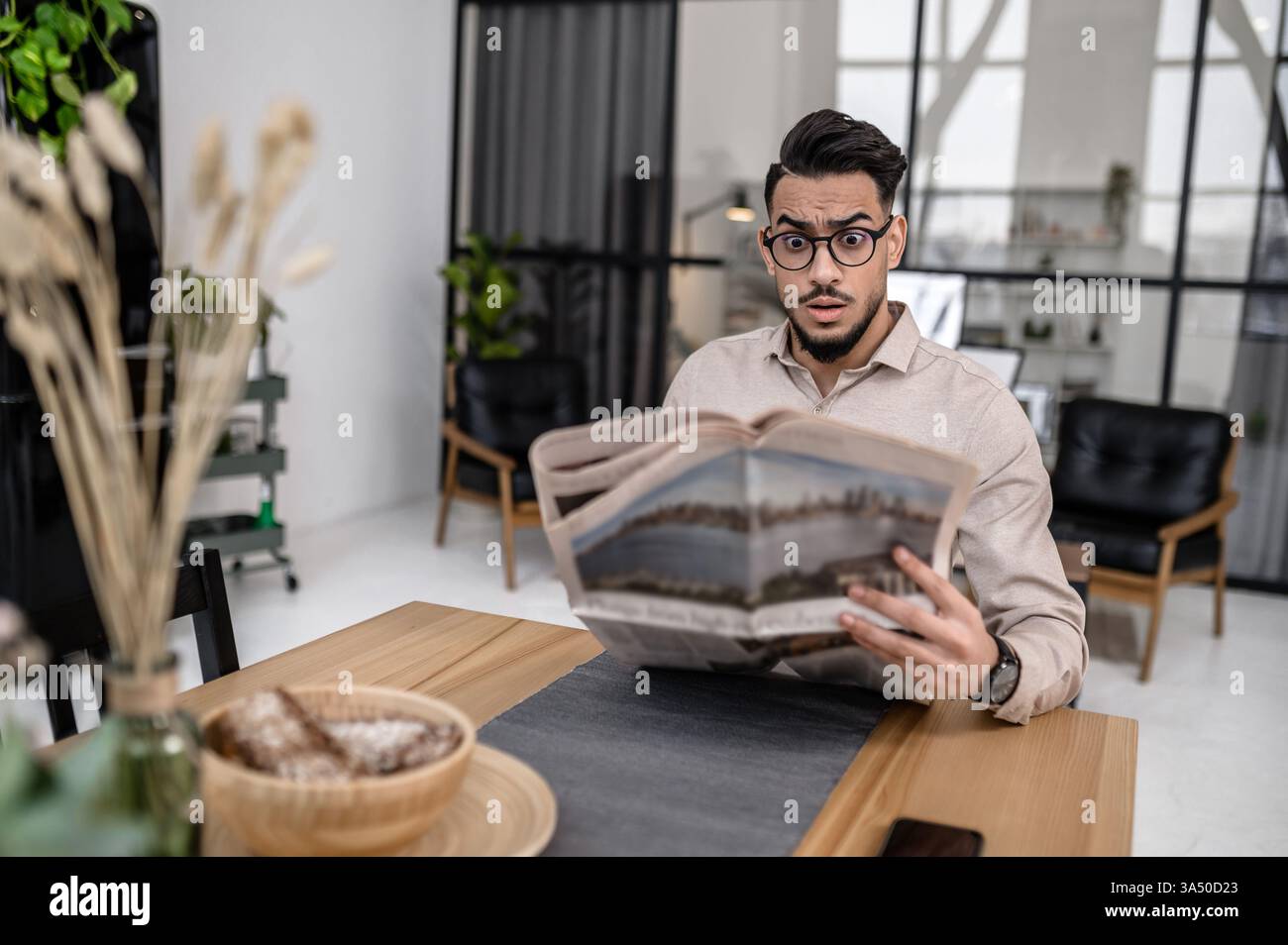Un homme arabe portant des lunettes montre une expression choquée en lisant à une table dans un salon. Le moment franc correspond au style de vie moderne et aux thèmes du bureau à domicile. Banque D'Images