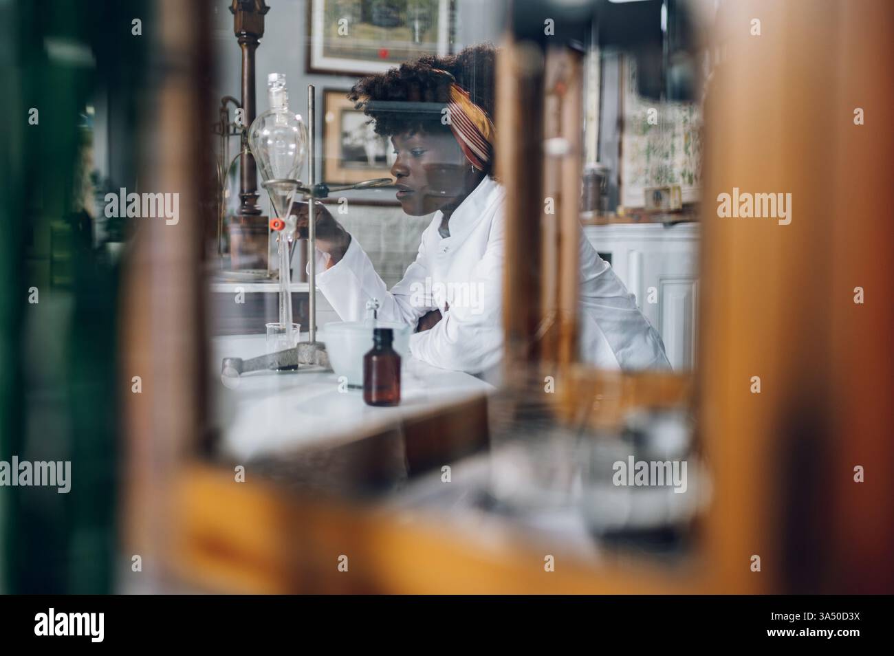 Vue à travers le verre d'un chimiste multiracial utilisant l'équipement de laboratoire pour préparer des médicaments dans une pharmacie vintage. Cette scène de santé authentique met en valeur la pharmacie, le patrimoine apothicaire et l’innovation pharmaceutique. Banque D'Images