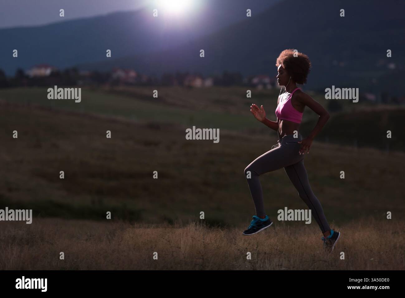 Femme noire avec afro dans les vêtements de fitness courant sur un champ au lever du soleil, écoutant de la musique avec des écouteurs. Une scène d'entraînement matinale énergique qui capture un style de vie actif et un entraînement en plein air. Idéal pour les campagnes de vêtements de sport, de bien-être et de fitness. Banque D'Images