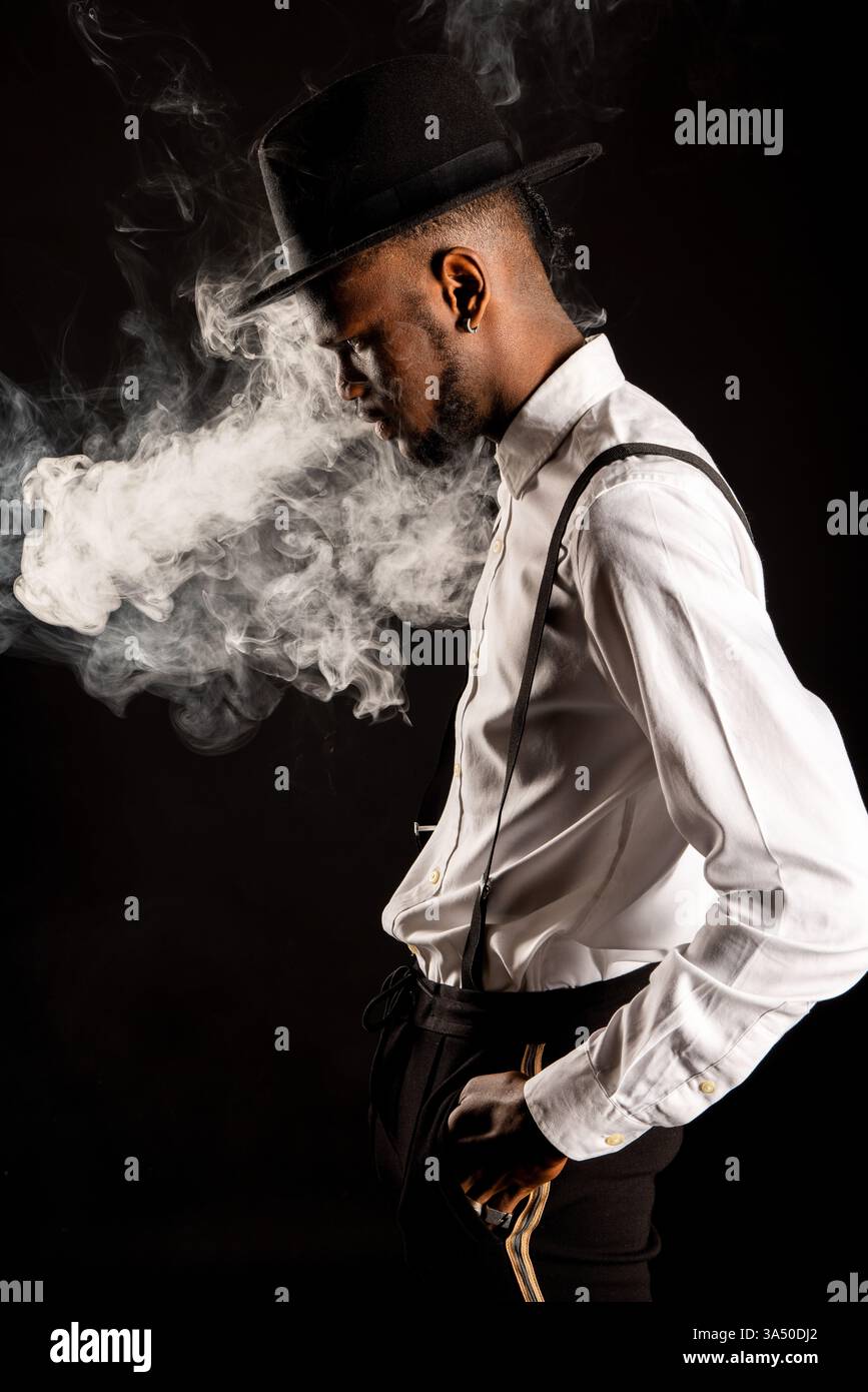 Photo studio d'un homme afro-américain exhalant la vapeur d'une e-cigarette sur un fond sombre. L’image du style de vie contemporain convient au vapotage, au marketing produit et aux contextes de mode urbaine. Idéal pour les campagnes explorant les tendances modernes et le style personnel. Banque D'Images