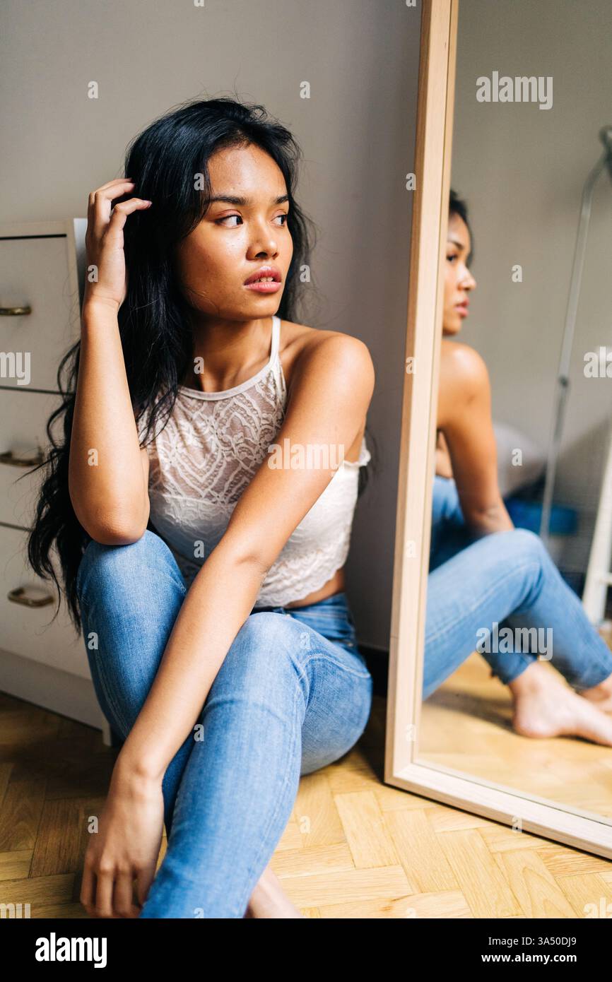 Femme asiatique assise sur le sol à la maison touche ses cheveux tout en regardant dans un miroir. Le moment intime et décontracté convient à la beauté, à la mode et aux thèmes de soins personnels. Idéal pour le style de vie à domicile et les contextes d'auto-soins. Banque D'Images