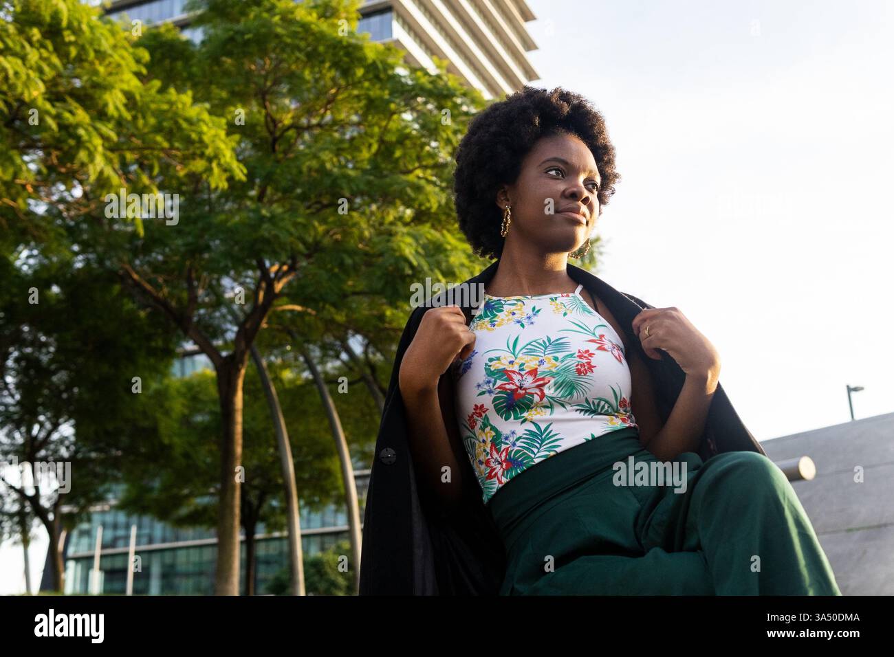 Photo en bas angle d'une jeune femme afro-américaine pensive avec une coiffure afro, assise sur un banc de parc dans un cadre urbain et ajustant sa veste. Ce portrait en plein air serein convient aux thèmes du mode de vie urbain, de la contemplation et du style personnel. Banque D'Images
