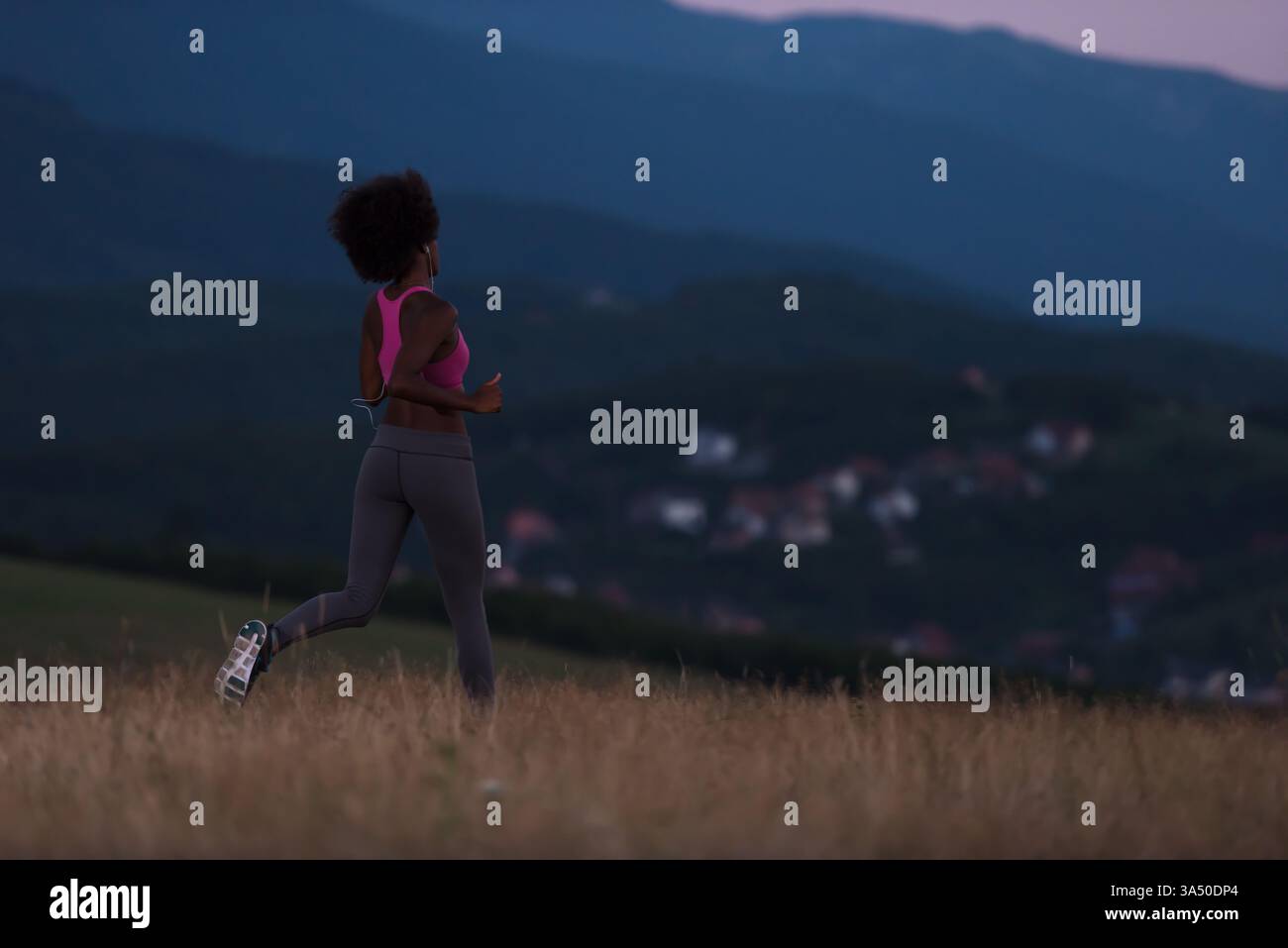 Jeune coureuse afro-américaine portant des écouteurs jogging dans un cadre naturel lors d'une belle nuit d'été. Ce moment de remise en forme en plein air capte le mouvement, l'énergie et la vitalité pour des histoires sur la course à pied et des modes de vie sains. Idéal pour les vêtements de fitness, l'entraînement d'endurance et les campagnes de bien-être. Banque D'Images