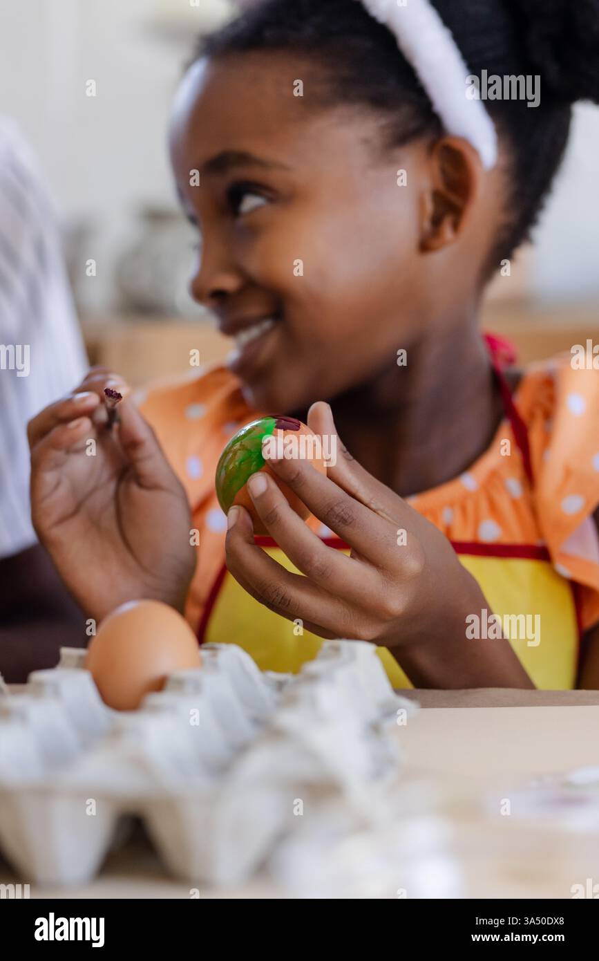 Fille afro-américaine peignant joyeusement l'oeuf de Pâques à la maison, portant un tablier coloré Banque D'Images