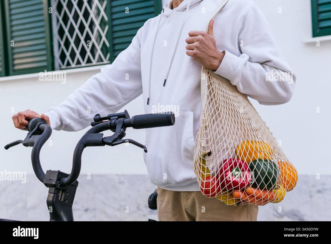 Un homme dans un pull porte un sac en maille rempli de fruits et légumes mûrs. Il se tient à côté d'un vélo dans une rue de la ville, suggérant des achats urbains et une alimentation saine. Idéal pour les thèmes écologiques, zéro déchet et de style de vie d'épicerie. Banque D'Images