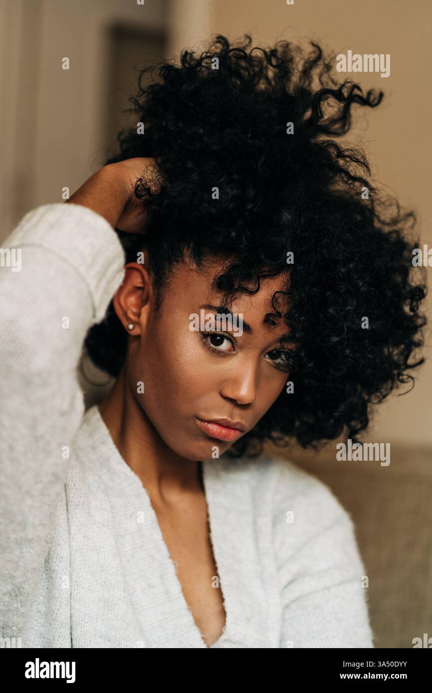 Portrait serein d'une femme noire se relaxant à la maison. Elle est assise sur un canapé, touchant ses cheveux dans un salon calme et confortable. Cette image fonctionne bien pour le style de vie et les caractéristiques de la vie domestique, soulignant le style intérieur et le bien-être personnel. Banque D'Images