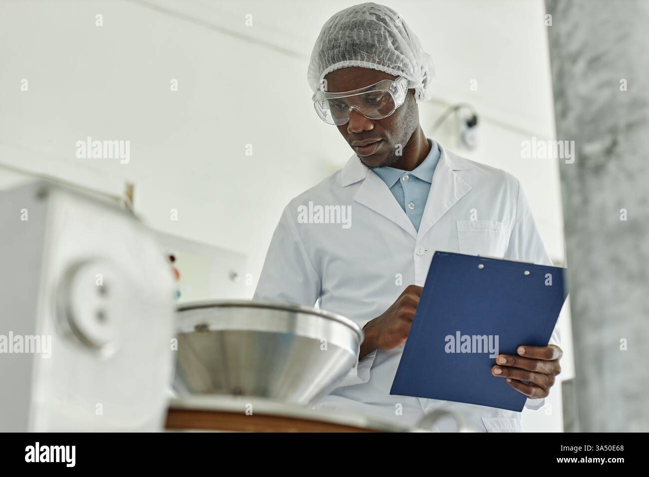 Portrait d'un jeune homme noir en blouse de laboratoire tenant une planche à pince, supervisant la production dans une usine alimentaire. Espace de copie disponible pour le marquage ou le message. Idéal pour l'industrie alimentaire, la fabrication ou l'imagerie sur le lieu de travail. Banque D'Images