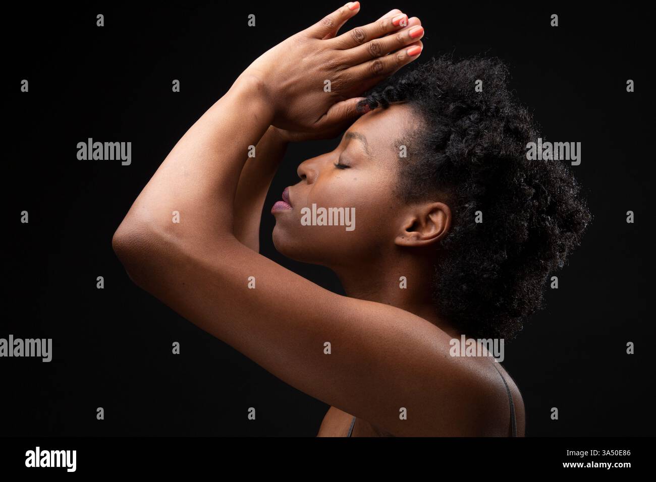 Portrait d'intérieur d'une femme afro-américaine aux cheveux naturels bouclés, les yeux fermés avec les bras levés dans la prière. Il capture la méditation, la santé mentale et les thèmes de soulagement du stress adaptés aux campagnes de bien-être. Banque D'Images