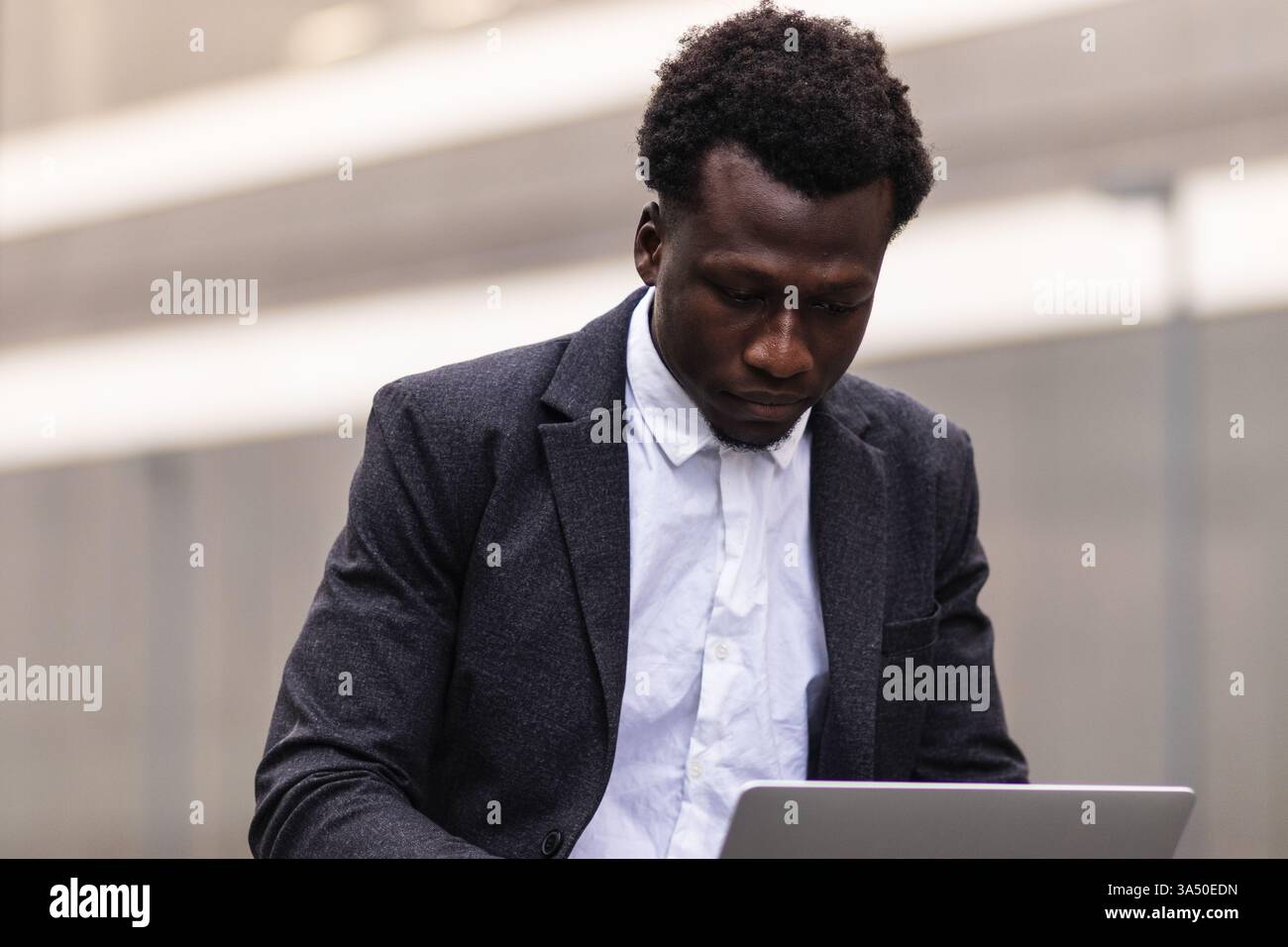 Homme d'affaires noir utilisant un ordinateur portable assis sur un banc dans la ville pendant la journée. Le cadre urbain extérieur met l'accent sur le travail moderne, la technologie et l'ambition professionnelle. Idéal pour les thèmes des affaires, de l'entrepreneuriat et de l'économie numérique. Banque D'Images