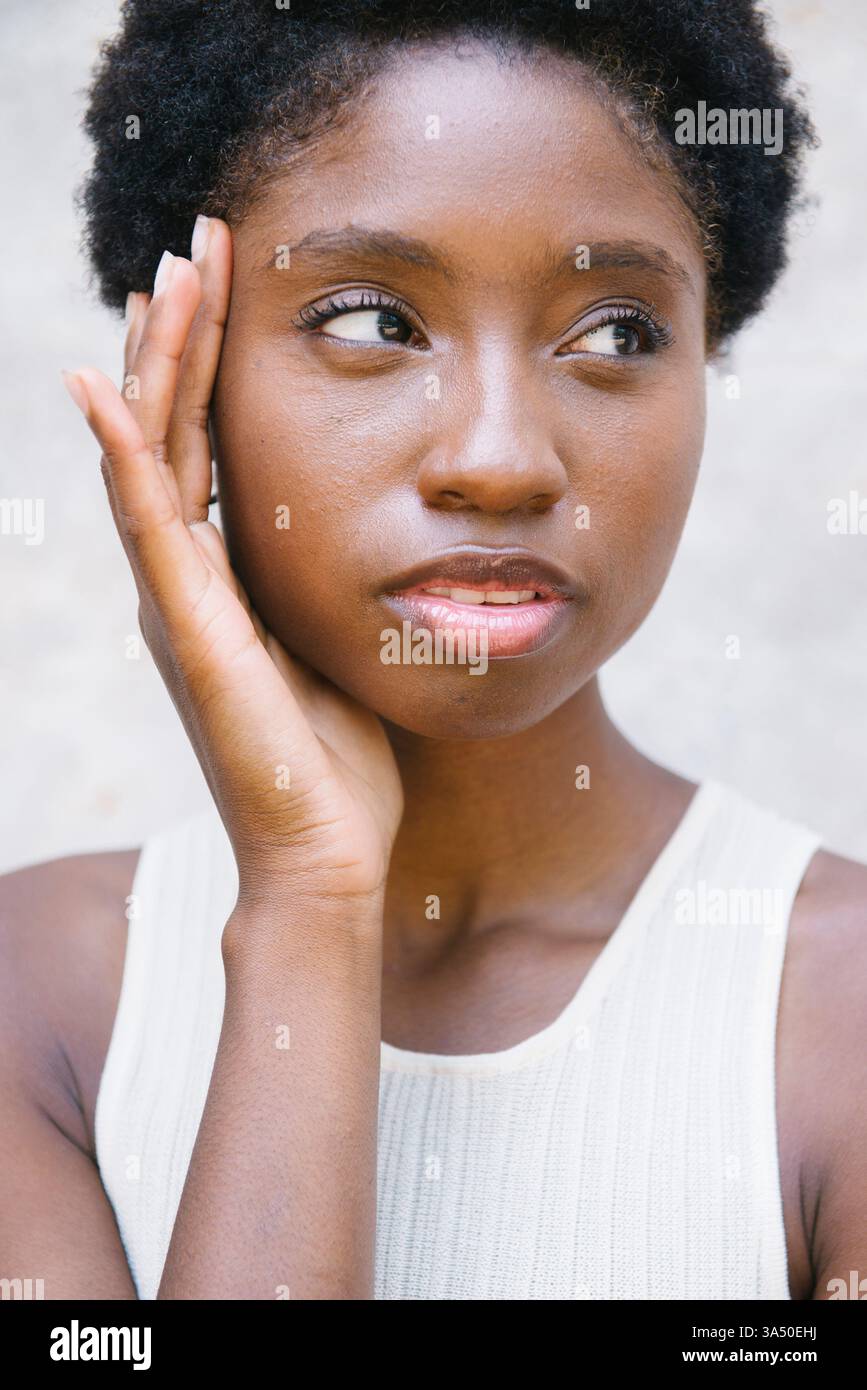Femme noire touchant son visage et regardant latéralement tout en se tenant debout contre un mur à la lumière du jour. La pose réfléchie communique la confiance et le style pour la beauté, la mode et les projets de bien-être personnel. Banque D'Images