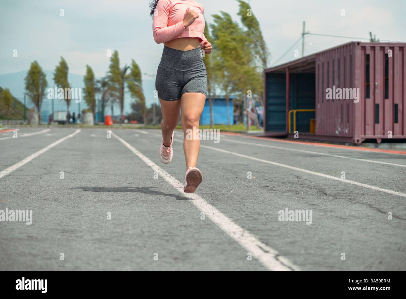 Athlète féminine non identifiable dans les jogging de vêtements athlétiques sur une piste de course de stade sur un matin nuageux. Idéal pour les graphismes de fitness, d'entraînement et de motivation avec un cliché recadré respectueux de l'intimité. La scène transmet l'énergie, la concentration et le bien-être extérieur. Banque D'Images