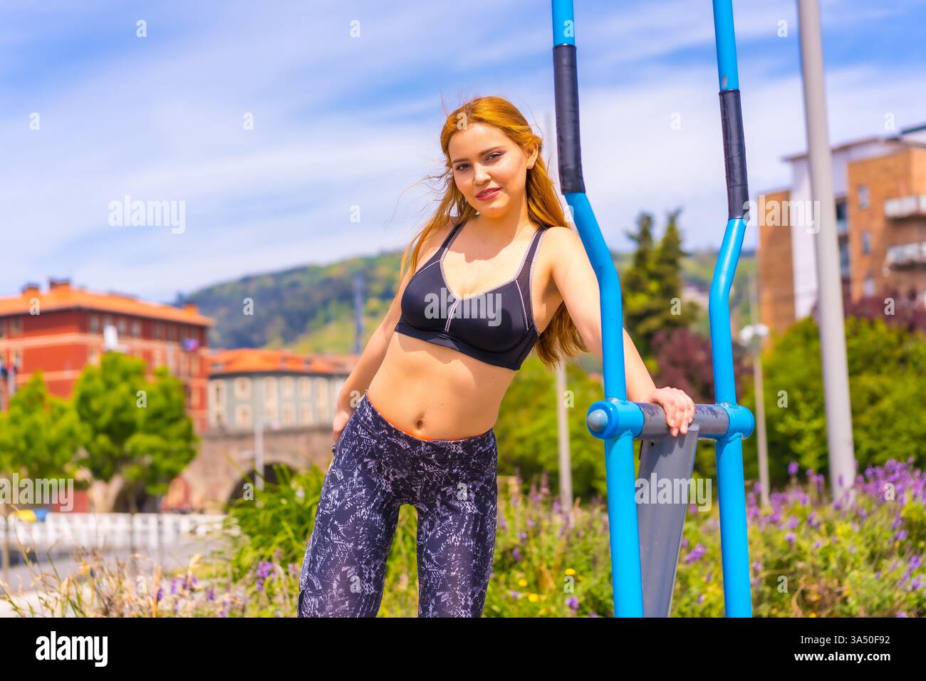 Une femme aux cheveux rouges en leggings violets et salopettes noires s'entraîne sur des machines de gymnastique dans un cadre urbain. La scène d'entraînement actif convient aux campagnes de fitness, de santé et de style de vie urbain. Banque D'Images